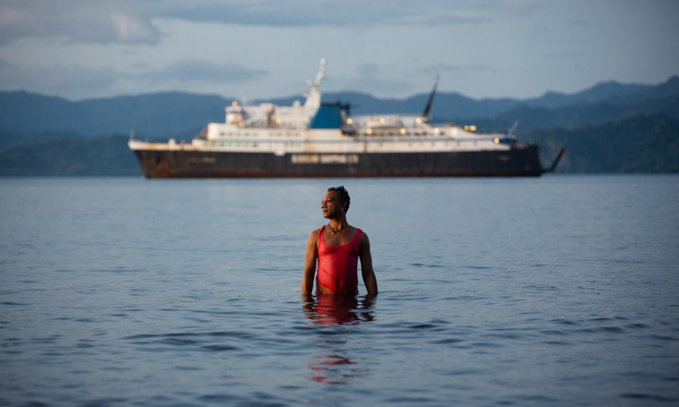 A man stands waist-deep in the sea in a red swimsuit, a ship behind him