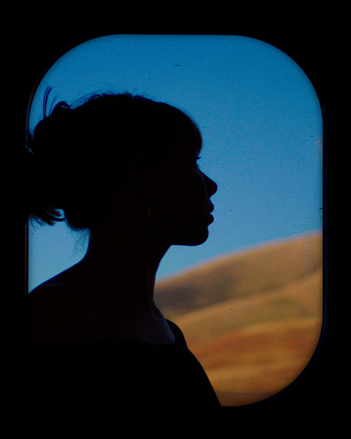 Silhouette of a woman in profile against a train window, blue sky and golden hills beyond